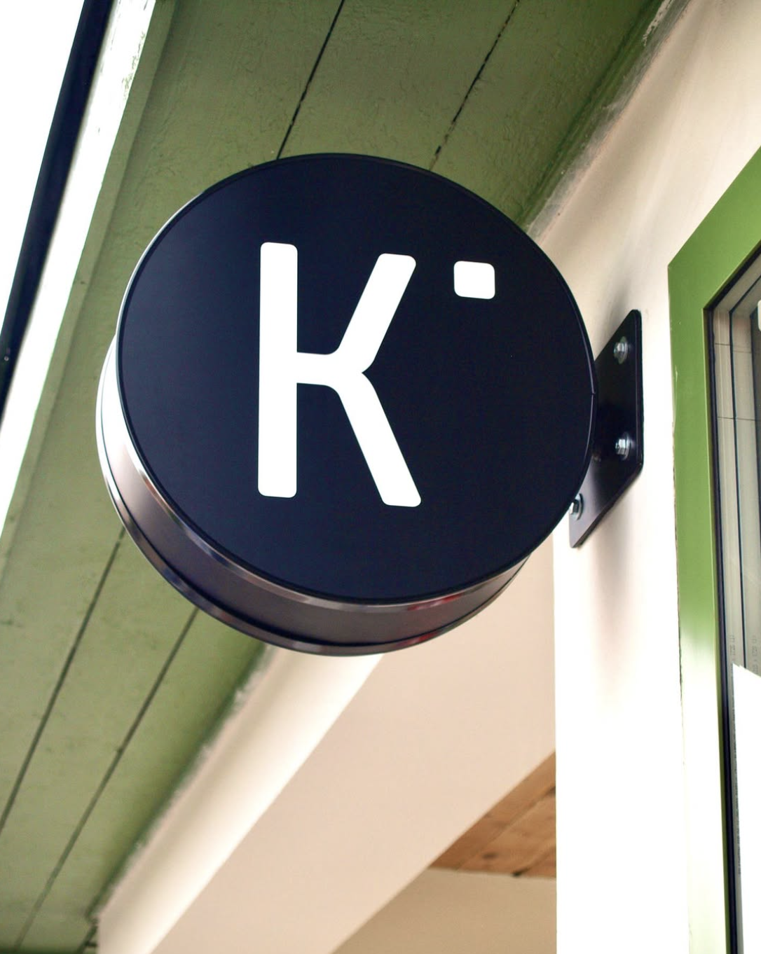 Kulatý navy cedník s logem K na zelené fasádě Kiosku